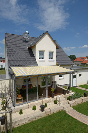 Modernes Einfamilienhaus mit Terrasse, Glasüberdachung und gelb-weißer Markise im Garten.