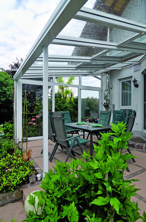 Überdachte Terrasse mit Glasdach, Gartenmöbeln und üppiger Bepflanzung im Sommer.