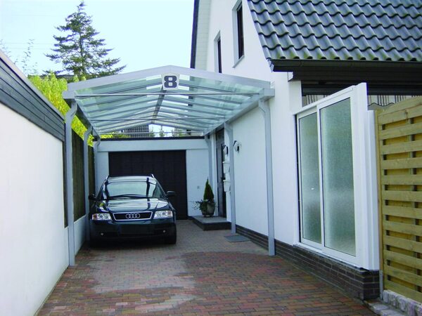 Carport mit Glasdach und parkendem Auto vor modernem Einfamilienhaus auf gepflasterter Einfahrt.
